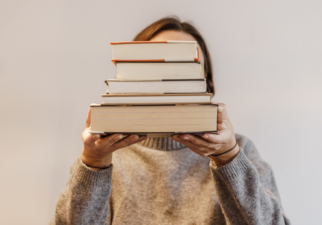 Person in Gray Sweater Carrying a Stack of Hardbound Books